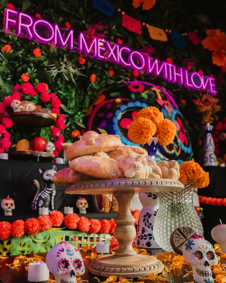 Pan de muerto on a wooden stand at a Día de Muertos altar, with marigolds, sugar skull candles and the From Mexico With Love neon sign